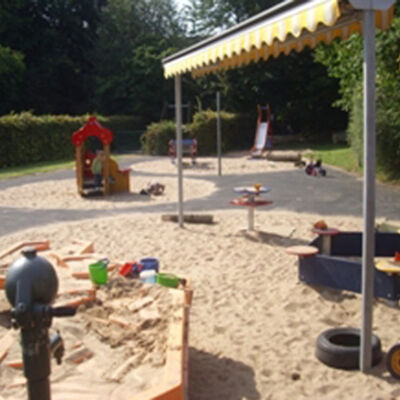 Bild vergrößern: Spielbereiche mit Wasserpumpe, Spielhaus, Rutsche und mit einem Sonnensegel berdachter Sandkasten.