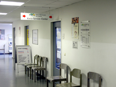 Bild vergrößern: Sthle stehen an einer Wand auf einem Gang mit einem Hinweisschild "Kreis-Service-Center".