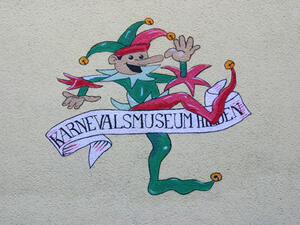 Bild vergrößern: Ein Narre schreitet über einen Banner mit der Aufschrift "Karnevalsmuseum Hilden".