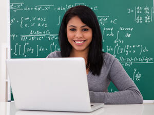 Bild vergrößern: Eine junge Frau sitzt an einem Laptop vor einer Tafel.