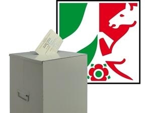 Bild vergrößern: Ein Stimmzettel steckt im Schlitz einer Wahlurne. Im Hintergrund befindet sich das Logo des Landes NRW.