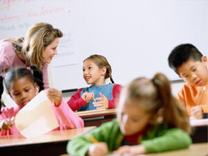 Bild vergrößern: Schulkinder sitzen an ihren Schultischen in einem Klassenraum. Eine Lehrerin steht am Tisch eines der Kinder.