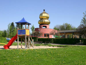 Bild vergrößern: Ein bunt bemaltes Klettergerüst mit Rutsche auf einem Spielplatz, im Hintergrund ein Gebäude mit goldener Kuppel.