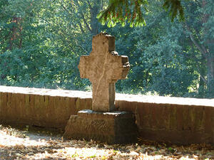 Bild vergrößern: Ein steinernes Kreuz steht auf einem Sockel vor einer kleinen Mauer umrandet von Bäumen.