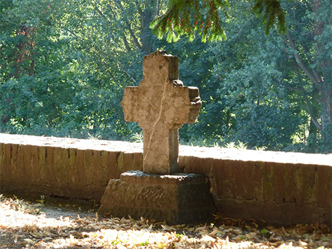 Bild vergrößern: Ein steinernes Kreuz steht auf einem Sockel vor einer kleinen Mauer umrandet von Bumen.