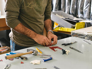 Bild vergrößern: Zwei Hände reparieren einen Gegenstand mit auf einem Tisch liegenden Werkzeug.