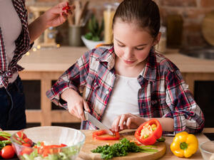 Bild vergrößern: Ein Mädchen sitzt an einem Tisch und schneidet Gemüse.