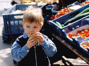 Bild vergrößern: Kleiner Junge mit Brötchen in der Hand steht an einem Marktstand.
