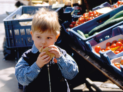 Bild vergrößern: Kleiner Junge mit Brtchen in der Hand steht an einem Marktstand.