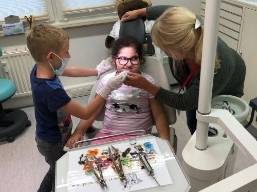 Bild vergrößern: Zwei Kinder mit Mundschutz untersuchen ein in einem Zahnarztstuhl sitzendes Mdchen.