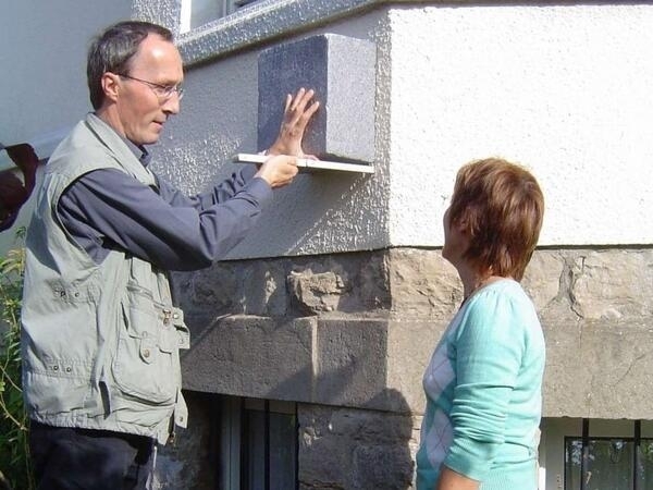 Bild vergrößern: Ein Mann misst mit einem Zollstock einen Stein an der Hauswand ab. Neben ihm steht eine Frau.