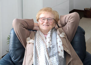 Bild vergrößern: Eine ältere Frau sitzt die Hände hinter dem Kopf verschränkt zurückgelehnt in einem Sessel.
