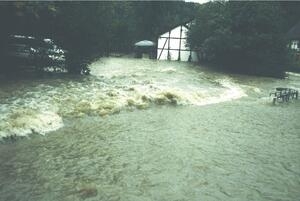 Bild vergrößern: Reißender Wasserstrom überschwemmt einen Straßenzug und Häuser.