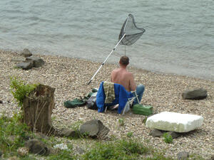 Bild vergrößern: Ein Angler sitzt am Wasser auf einem Stuhl, neben sich eine Angel und ein Fangnetz.