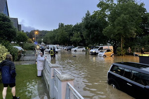 Bild vergr&ouml;&szlig;ern: Feuerwehr im Einsatz auf einer kniehoch &uuml;berfluteten Stra&szlig;e in Erkrath. Die Einsatzkr&auml;fte besprechen sich mit den Anwohnern. Eine Dame steht im Regenmantel an ihrem Gartenzaun im Vorgarten. Der Zaun steht auf einer kleinen Mauer, die das Wasser zur&uuml;ckh&auml;lt.