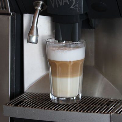 Bild vergrößern: Ein gefülltes Glas Milchkaffe mit aufgeschäumter Milch steht unter dem Abfüllhahn einer Kaffeemaschine.