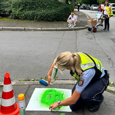 Bild vergrößern: Polizistin die mit hilfe einer Schablone grüne Fußspuren auf einen Gehweg sprüht.