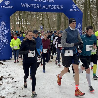 Bild vergrößern: Läufer starten bei der Hildener Winterlaufserie unter einem blauen Bogen durch eine matschige und schneebedeckte Landschaft.