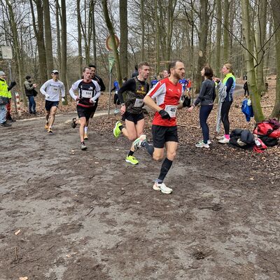 Bild vergrößern: Mehrere Teilnehmer in Sportklediung laufen auf einem matschigen Waldweg bei der Hildener Winterlaufserie, mit Zuaschauern am Rand des Weges.